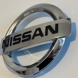 Peut inclure: Un emblème de voiture chromé avec le mot "NISSAN" en lettres noires.