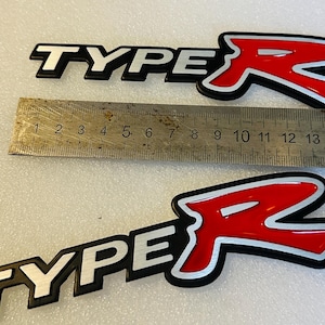 Peut inclure: Deux emblèmes "TYPE R" noirs et blancs avec un "R" rouge dans une police de caractères stylisée. Les emblèmes sont sur une surface blanche avec une règle pour l'échelle.
