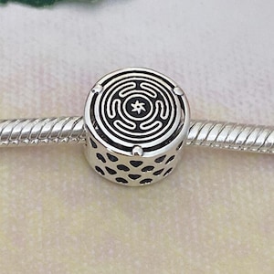 May include: A silver charm bracelet with a cylindrical charm featuring a maze design. The charm has a black center with a star-like symbol and heart-shaped cutouts. The words "Modern Dreamy" are in the upper left corner.