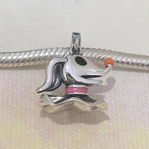 Könnte beinhalten: Ein silberner Charm-Anhänger mit Zero aus The Nightmare Before Christmas. Der Anhänger hat einen weißen Kopf mit schwarzen Augen, einen rosa Kragen und eine orangefarbene Nase. Der Anhänger ist an einem silbernen Armband befestigt.