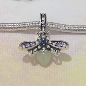 May include: A silver charm in the shape of a bee, with purple and blue accents. The bee charm has a light green, heart-shaped body and is suspended from a silver chain. The charm is detailed and delicate.