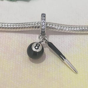 Op de afbeelding: Een zilveren bedelarmband met een bedel met een zwarte achtbal en een zilveren keu. De bedel is bevestigd aan een zilveren ring met heldere stenen. De tekst "Modern Dreamy" staat linksboven.