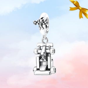 Könnte beinhalten: Ein silberner Charm-Anhänger mit einem Formel-1-Rennwagen-Design. Der Anhänger hat ein detailliertes, dreidimensionales Aussehen mit sichtbaren Rädern, einem Cockpit und einem Frontflügel. Der Anhänger hängt an einem runden, dekorativen Element mit einem karierten Flaggen-Design.