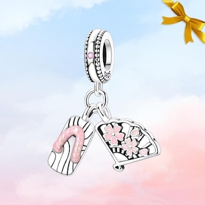 May include: A silver charm with a pink and white sandal and fan design. The charm features a pink flower and sandal strap, with a decorative bead at the top. The charm is suspended from a silver ring.