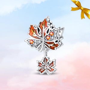 May include: A silver charm featuring a large maple leaf with orange enamel, clear cubic zirconia stones, and orange accent gems. A smaller, matching maple leaf charm hangs from the bottom of the larger leaf, both crafted with detailed silver veins.