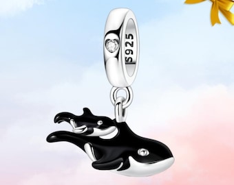 May include: A silver charm with a black and white orca whale design. The charm features a small, clear gemstone and is stamped with "S925". The orca whale has a baby orca whale attached to it. The charm is suspended from a silver loop.