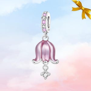 May include: A silver charm featuring a pink bell flower design. The charm has a ring set with pink and clear stones. A clear, sparkling stone hangs below the flower. The background is a pink and blue gradient.