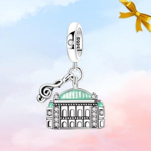 Può includere: Un charm in argento con un design a forma di edificio e una chiave di violino. L'edificio ha un tetto verde menta ed è decorato con cristalli trasparenti. Il charm è contrassegnato con "S925" e un piccolo cristallo. Lo sfondo è un gradiente di rosa e blu.