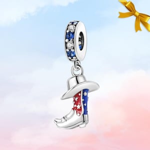 May include: A silver charm featuring a cowboy boot and hat. The boot is decorated with red and blue enamel and white stars, while the hat is white. The charm's top has blue and clear crystal accents, suitable for a bracelet or necklace.