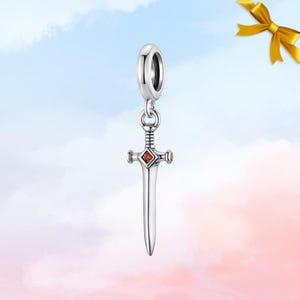 Könnte beinhalten: Ein silberner Schwert-Charm mit einem roten Edelstein in der Mitte des Griffs. Der Charm hat oben eine Schlaufe zum Befestigen an einem Armband oder einer Halskette. Der Schwert-Charm ist ein Anhänger.