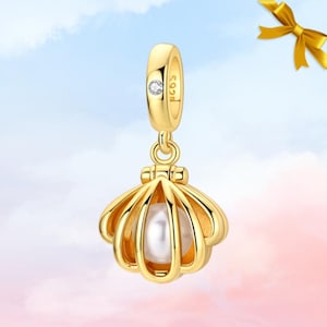 May include: A gold-colored charm pendant featuring a seashell design with a pearl inside. The pendant has a small diamond accent and is suspended from a loop. The background is a gradient of blue and pink.