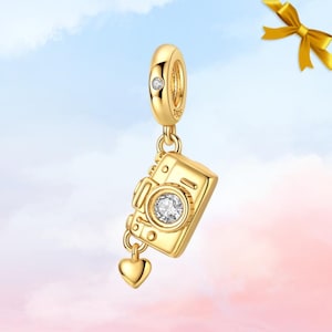 Peut inclure: Un charm doré en forme d'appareil photo. Le charm comprend un petit pendentif en forme de cœur et une gemme étincelante au centre de l'objectif. Le charm est suspendu à un anneau doré avec une petite gemme.