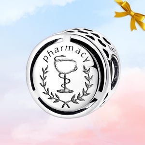 May include: A silver charm with the word "pharmacy" encircling a caduceus symbol within a laurel wreath. The charm has a detailed design and is set against a soft, colorful background.