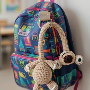 May include: A blue backpack with a colorful pattern and pink accents hangs on a hook. A crocheted, light brown sloth-shaped keychain is attached to the backpack's handle. The sloth has white accents and a brown nose.
