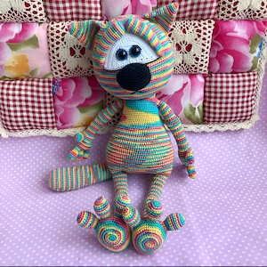 May include: A handmade, colourful crochet cat doll with a striped body and tail. The cat has a white face, blue eyes, and a black nose. The doll is sitting on a purple polka dot surface, with a patchwork background.
