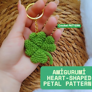 Könnte beinhalten: Ein grüner, gehäkelter vierblättriger Kleeblatt-Schlüsselanhänger mit einem goldfarbenen Ring und einer Kette. Der Schlüsselanhänger trägt den Text "Crochet PATTERN" und "AMIGURUMI HEART-SHAPED PETAL PATTERN" in einer grünen Box.