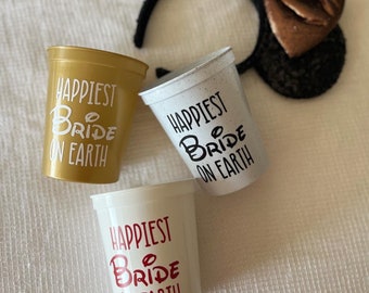 May include: Three plastic cups with the text "Happiest Bride on Earth" in different colors. The cups are white, gold, and red.
