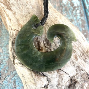 May include: A green jade pendant in a hook shape, suspended from a black cord. The pendant has a smooth, polished surface with variations in color. The cord is knotted at the top. The pendant is displayed on a piece of driftwood.