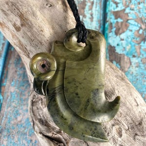 May include: A greenstone pendant carved in a traditional hook shape, suspended from a black cord. The pendant features a swirling design and a small, round accent with iridescent details. The background is a weathered wooden surface.