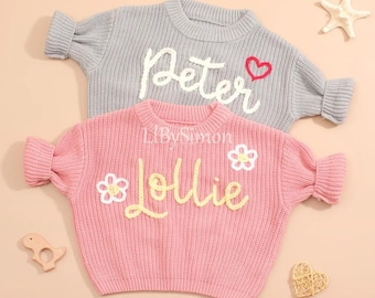 May include: Two knitted sweaters with embroidered names. The pink sweater has the name "Lollie" embroidered in yellow with white flowers. The gray sweater has the name "Peter" embroidered in white with a red heart.