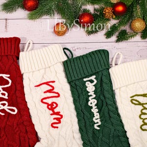 May include: Four cable-knit Christmas stockings in red, white, and green, each with a different name embroidered in a contrasting colour. Festive holiday decor with evergreen branches and baubles.
