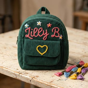 May include: A dark green corduroy backpack featuring the embroidered name "Lilly B." in pink, accented with small colourful embroidered flowers and a yellow embroidered heart on the front pocket, accompanied by several colourful embroidery thread skeins.