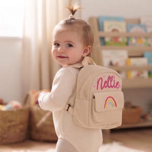 May include: A beige corduroy mini backpack featuring the name Nellie embroidered in pink script with small pink hearts above it and a colorful rainbow graphic stitched onto the front pocket, complete with adjustable shoulder straps.