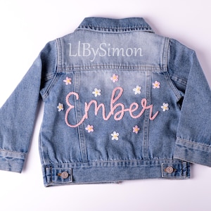 May include: A blue denim jacket with pink embroidery of the name "Ember" and white flowers. The text "LLBySimon" is embroidered on the back of the jacket.
