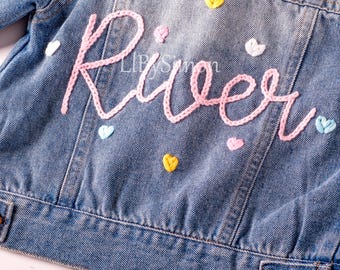 Könnte beinhalten: Eine blaue Jeansjacke mit dem Namen "River", der in rosa Garn gestickt ist. Die Jacke ist mit bunten Herzen verziert.
