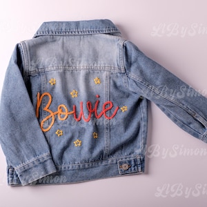 May include: A light-wash denim jacket with the name "Bowie" embroidered in orange and red cursive script on the back. Small yellow star accents surround the name. The jacket has a classic collar and button closures.