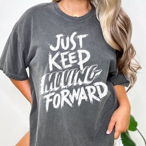 Pode incluir: Uma camiseta cinza-chumbo oversized com um gráfico branco que diz "Just Keep Moving Forward".