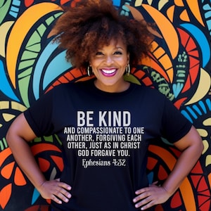 Puede incluir: Una mujer lleva una camiseta negra con un gráfico blanco que dice "BE KIND AND COMPASSIONATE TO ONE ANOTHER, FORGIVING EACH OTHER, JUST AS IN CHRIST GOD FORGAVE YOU. Ephesians 4:32". Está de pie frente a una pared colorida con diseños abstractos.