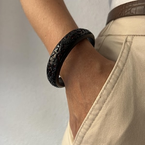 Può includere: Un bracciale rigido circolare di colore marrone scuro caratterizzato da un intricato motivo floreale intagliato su tutta la superficie. Il bracciale presenta una finitura liscia e lucida e una struttura solida e continua che avvolge il polso.