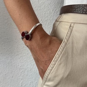 Op de afbeelding: Een zilveren armband met twee ronde, dieprode cabochonstenen die in getextureerde metalen zettingen aan beide open uiteinden zijn geplaatst. De armband wordt gedragen om een pols bij een beige broek met een donkerbruine riem met textuur.