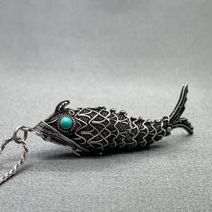 May include: A silver-toned fish pendant featuring intricate filigree scale patterns and a single round, bright turquoise stone set as the eye. The pendant is attached to a twisted silver-toned metal chain link, showcasing detailed metalwork.