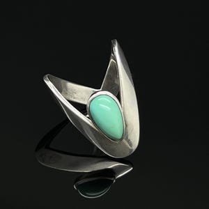 Op de afbeelding: Een zilveren ring met een gladde, druppelvormige turkoois steen gezet in een gepolijste, V-vormige metalen band. De zilveren band heeft een hoogglans afwerking en een puntig, architectonisch ontwerp dat de centrale lichtblauw-groene steen omlijst.