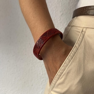 Può includere: Un bracciale rigido rosso scuro caratterizzato da un motivo geometrico ripetitivo di semicerchi concentrici. Il bracciale presenta una finitura opaca liscia e una struttura a fascia larga e rigida che circonda il polso, mostrando un design ad arco.