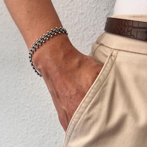 Könnte beinhalten: Ein silbernes Kettenarmband mit einem sich wiederholenden Muster aus kleinen, miteinander verbundenen kreisförmigen Gliedern mit winzigen kugelförmigen Perlen an jeder Verbindungsstelle, getragen am Handgelenk über einer beigen Hose mit einem dunkelbraunen Ledergürtel.