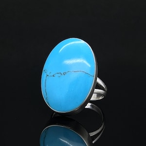 Peut inclure: Une bague en argent ornée d'une grande pierre turquoise ovale bleu vif avec de fines veines sombres. La pierre est sertie dans une monture en argent lisse, et l'anneau présente un design à double branche qui s'affine vers l'arrière de la bague.
