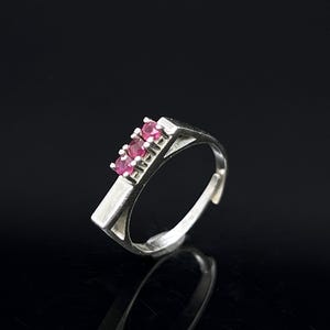 Peut inclure: Une bague argentée ornée de trois petites pierres rondes à facettes de couleur rouge rosé, disposées en ligne. L'anneau présente un design géométrique à cadre ouvert avec une partie supérieure plate et large qui s'affine vers le bas.
