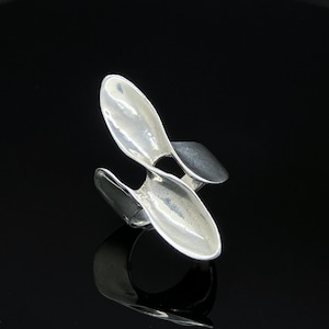 Può includere: Un anello in argento dal design scultoreo aperto, caratterizzato da due forme concave allungate simili a petali che si curvano verso l'esterno dalla fascia centrale. La superficie metallica presenta una finitura lucida e riflettente.