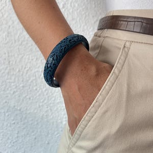 Pode incluir: Uma pulseira rígida azul-escura com um padrão detalhado de flores e folhas em relevo esculpido em toda a sua superfície. A pulseira tem um perfil arredondado e suave com um acabamento mate, envolvendo o pulso com uma estrutura sólida.