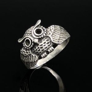 May include: A silver-toned ring featuring a detailed owl design on the front. The owl has large circular eyes, textured feathers on its wings, and a patterned chest. The band of the ring is smooth and polished, contrasting with the intricate metalwork.
