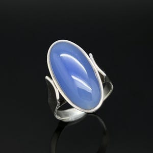 Può includere: Un anello in argento con una grande pietra blu ovale e liscia a cabochon, incastonata in una montatura in metallo lucido. La fascia presenta un design diviso con dettagli laterali testurizzati che si curvano verso l'alto per sostenere la pietra.