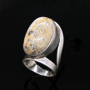 May include: A silver ring featuring a large, oval-shaped stone with a complex, swirling pattern in shades of cream, tan, brown, and grey. The stone is set in a smooth, polished silver bezel, mounted on a wide, open-style silver band.