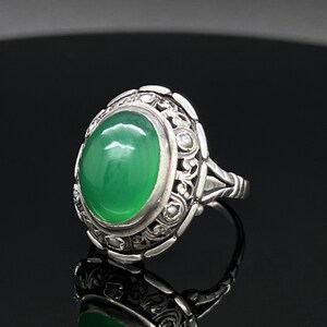 Könnte beinhalten: Ein Silberring mit einem großen, ovalen, im Cabochon-Schliff geschliffenen grünen Stein in der Mitte. Die Metallschiene und die Fassung sind mit komplizierten, erhabenen Schnörkelmustern und kleinen, runden, weißen Akzenten verziert.