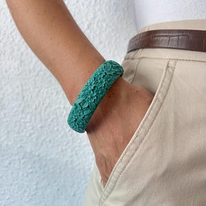 Może przedstawiać: Turkusowa bransoletka typu bangle z wygrawerowanym, szczegółowym i ciągłym wzorem kwiatowym, składającym się z małych kwiatów i liści pokrywających całą zewnętrzną powierzchnię sztywnej, okrągłej obręczy, co nadaje akcesorium teksturowany wygląd.