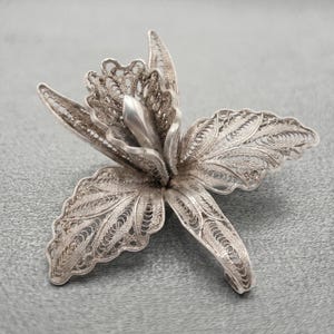 May include: A silver brooch shaped like a flower, featuring intricate filigree wirework on the petals and a smooth, solid silver center. The metallic piece displays a detailed, open-work pattern throughout the leaves, creating a delicate floral design.