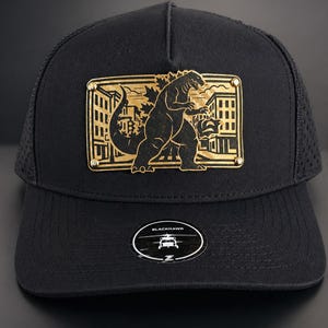 May include: Black baseball cap with a gold-colored Godzilla design on the front. The cap has a mesh back and a curved brim. The Blackhawk logo is on the front.