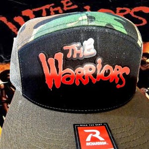 May include: A trucker hat with a black front panel displaying the words "The Warriors" in red and white lettering. The top of the hat has a camouflage pattern, and the brim is a dark olive green. The hat has a mesh back and a Richardson tag.